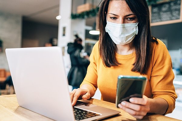  Ženska z zaščitno masko na obrazu sedi pri mizi in dela na prenosni računalnik. V eni roki drži mobilni telefon s katerega bere.